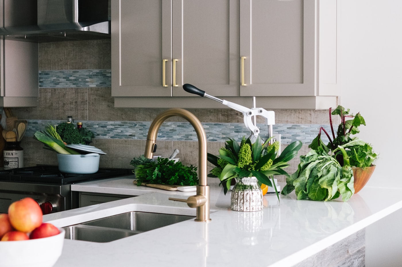 A kitchen counter with herbs and vegetables, warm cabinets and a brass tap
